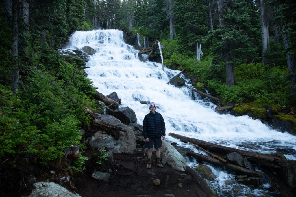 A hiker poses in front of a waterfall on the hike. 