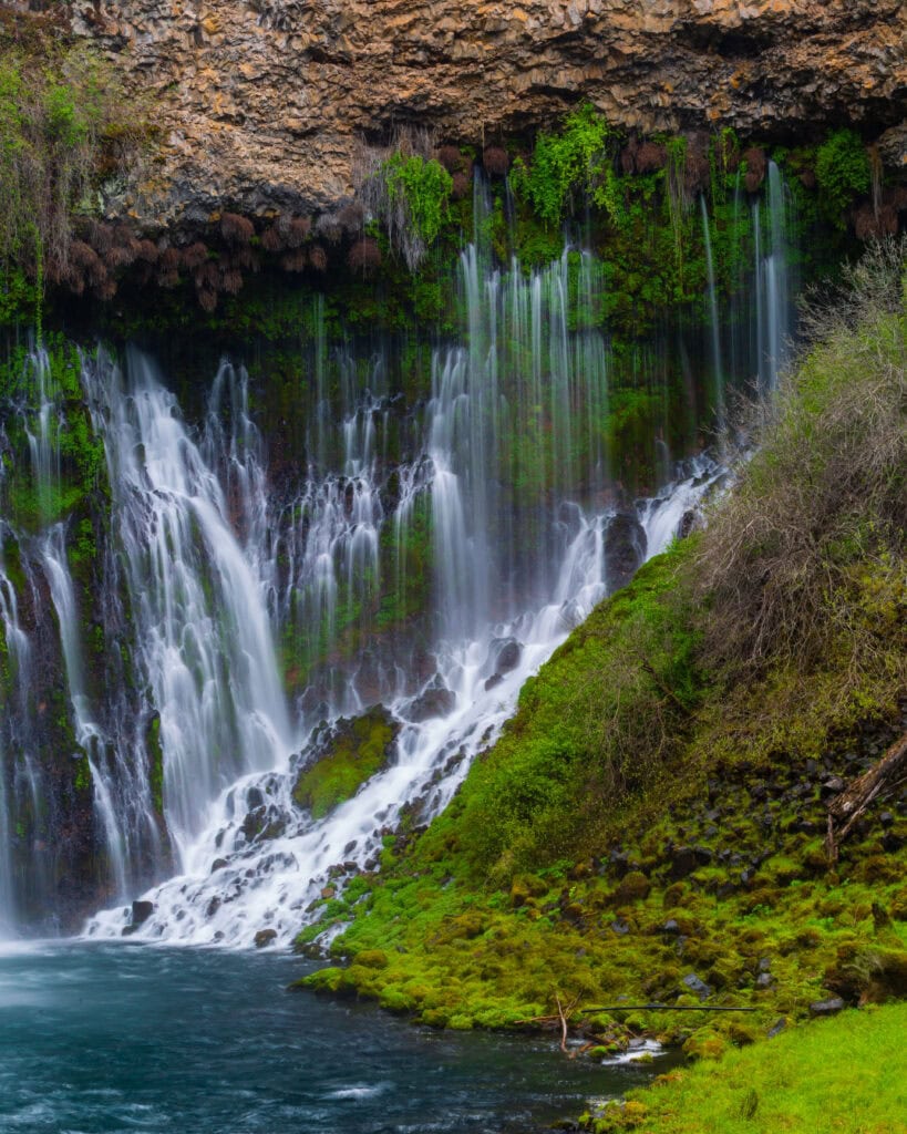 Burney Falls dropping into a calm teal pool framed by green vegetation at McArthur-Burney Falls Memorial State Park, California