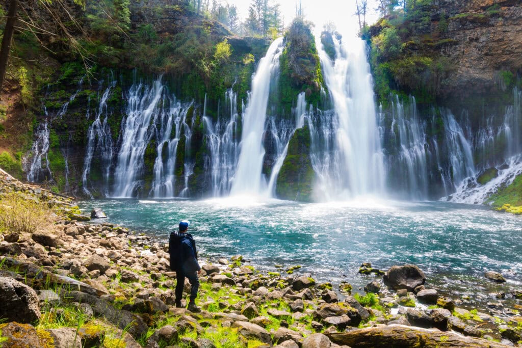 Hiker standing at the base of Burney Falls with the wide cascade filling the frame at McArthur-Burney Falls Memorial State Park, California