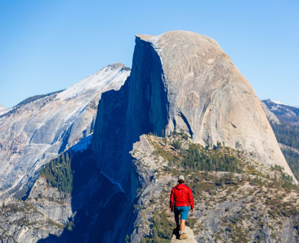 a tourist looks out at halfdome after hiking the four mile trail in yosemite. he's wearing a red jacket