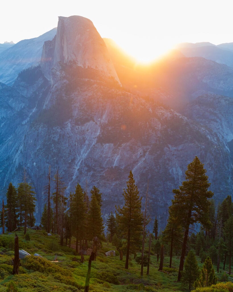 the sun rising over half dome with trees in the foreground