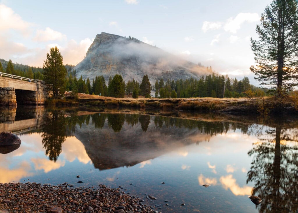 a foggy morning looking at lembert dome in yosemite national park with a river running through in the foreground with clear reflections of trees on the water