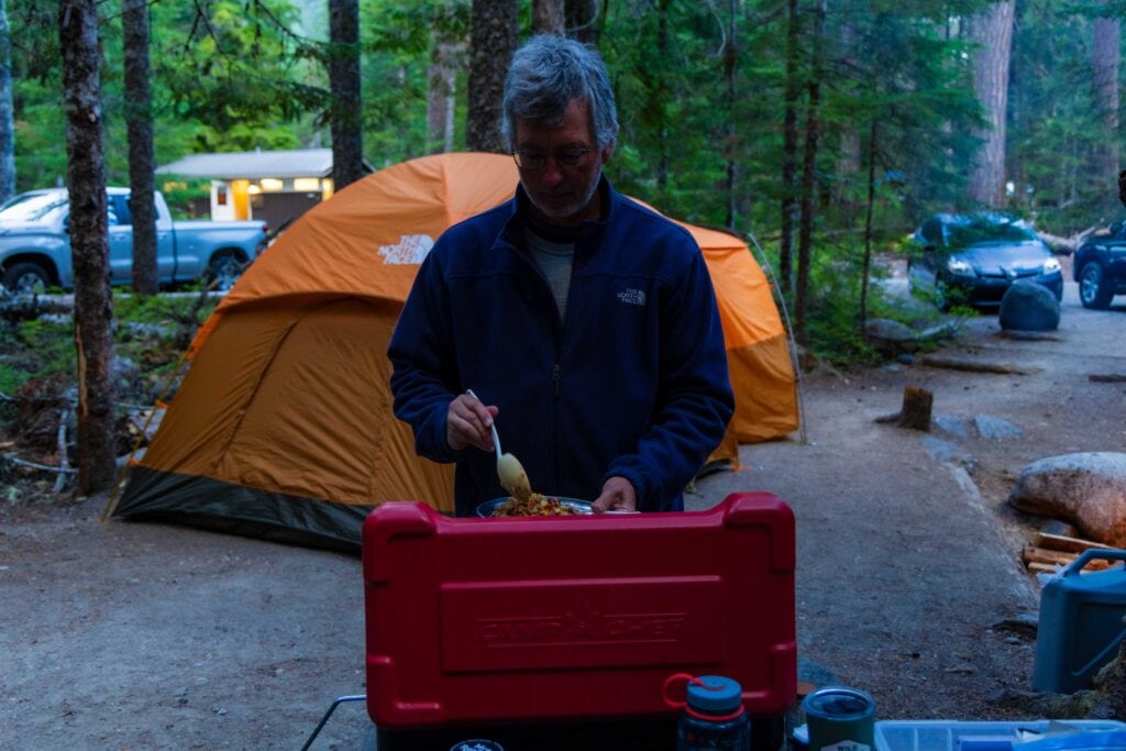 A male stands near a camp stove serving food.