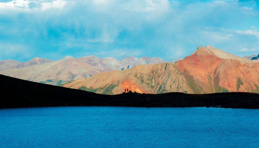 two people stand on a ridgeline across a lake with mountains behind them