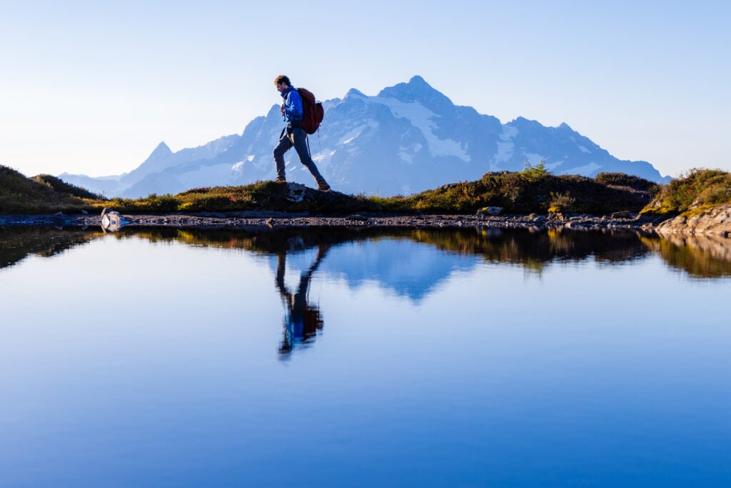 A hiker walks with a reflection of him and a mountain in the water below.