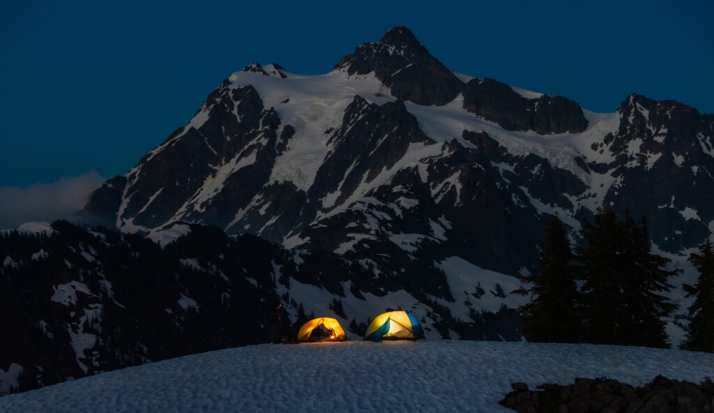 two tents sitting on the snow being illuminated at night with a snow mountain behind it