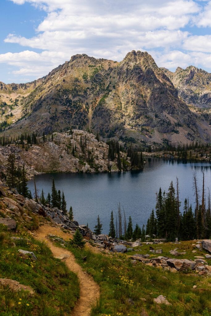 the trail cuts through the green grasses with a lake and mountains and clouds above