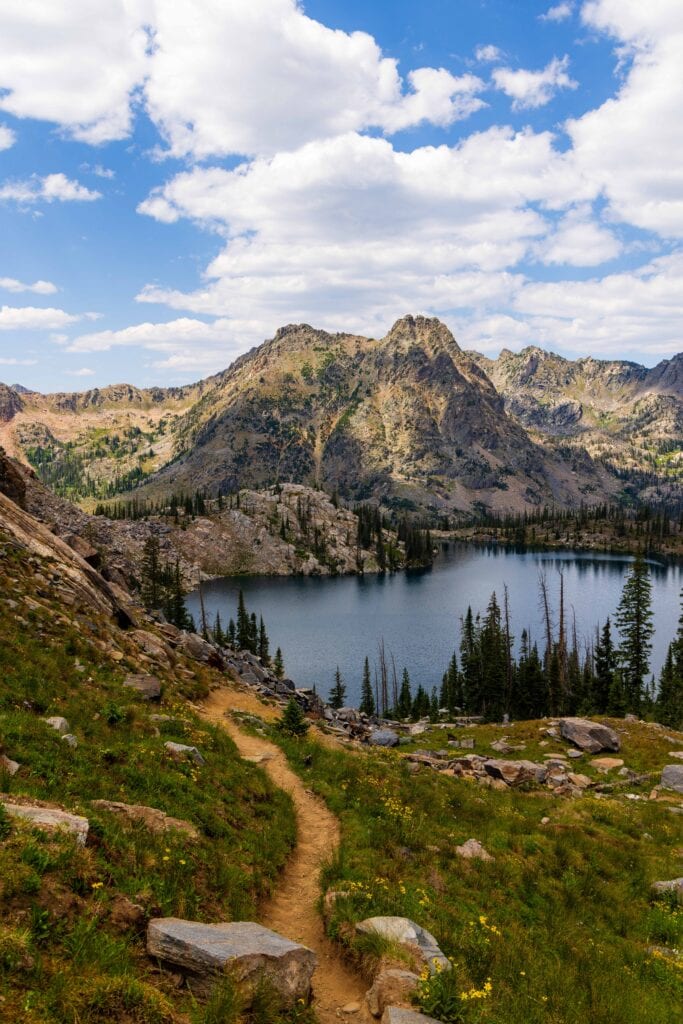 the trail cuts through the green grasses with a lake and mountains and clouds above