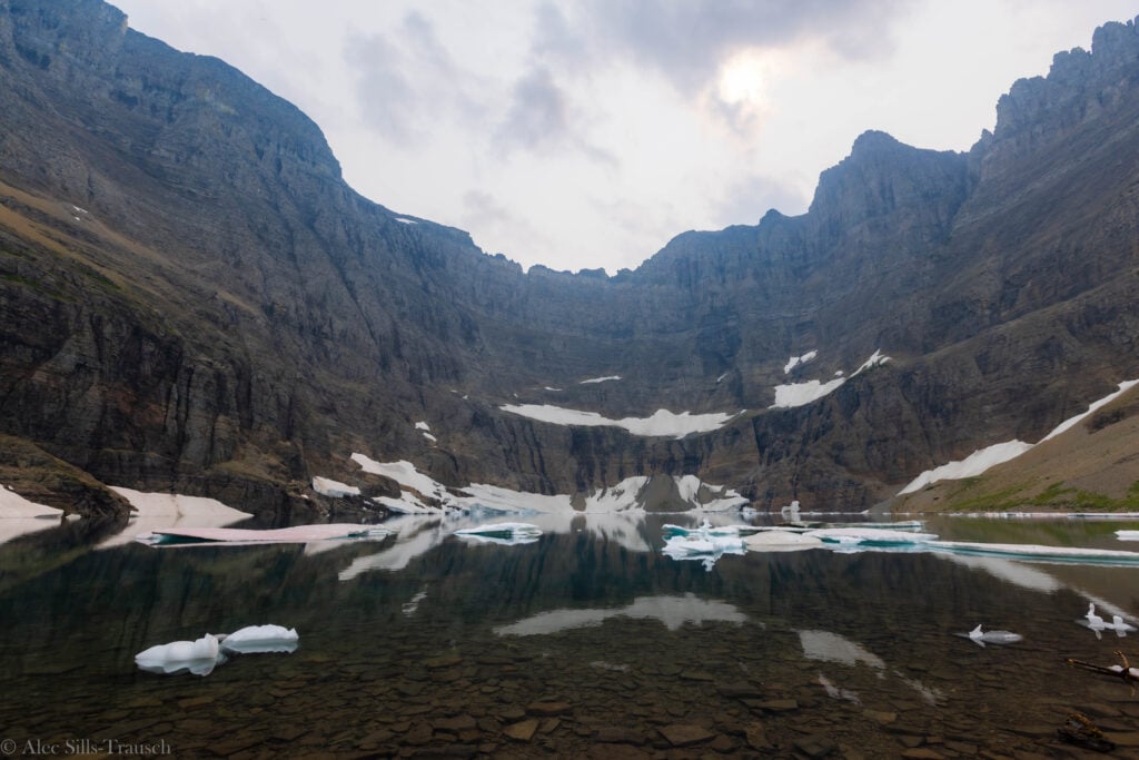 a massive roclwall with an alpine lake below and glacier floating in it