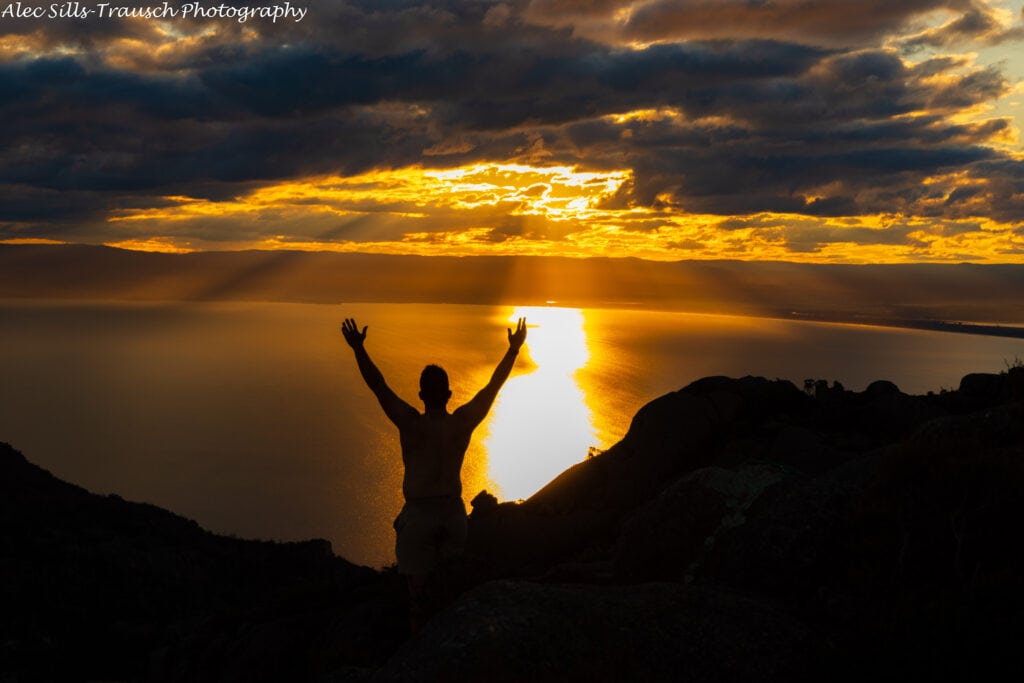 A hiker raises his hands in front of the sunset glow. 