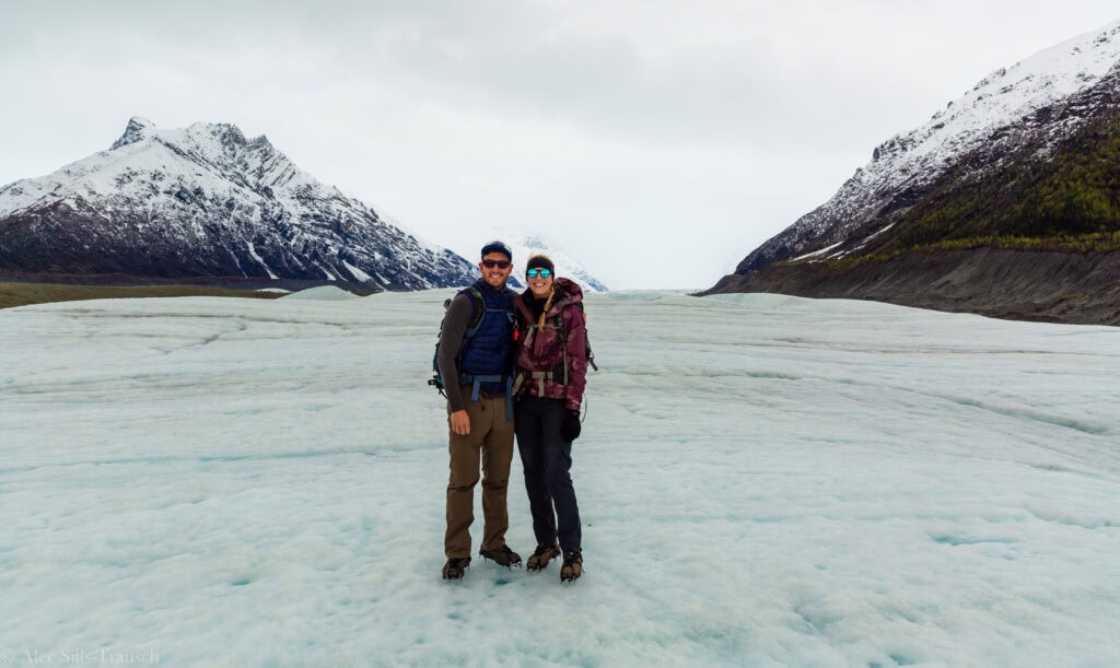 two people on a glacier in alaska with snowy mountains behind them