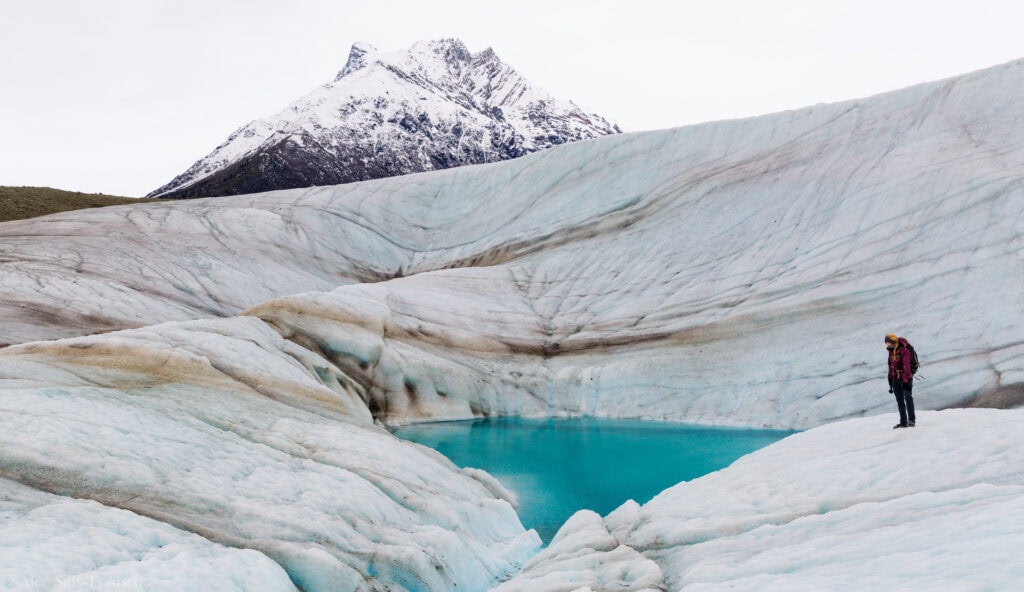 A hiker stands on a glacier in alaska in front of a pool