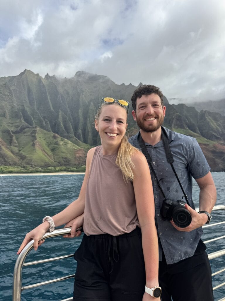 two travelers smile at the camera with the na pali coast in the background
