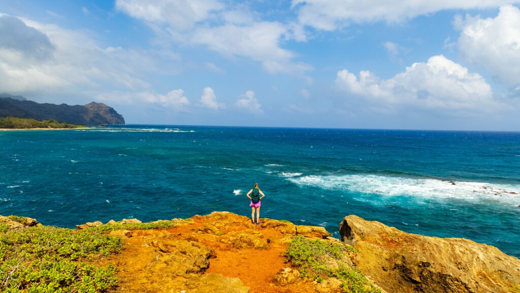 a woman stands on the edge of a cliff overlooking the pacific ocean in kauai on the Maha'ulepu Heritage Trail