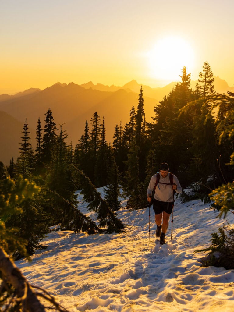 a male hiker walks towards the camera with hiking poles and the sun above him