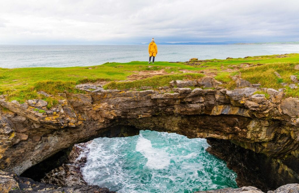 a tourist in a yellow jacket looks out at the ocean standing on a fairy bridge