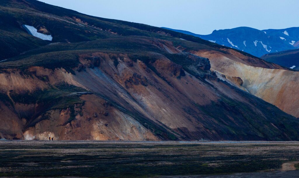 two people far off walk next to a mountain side