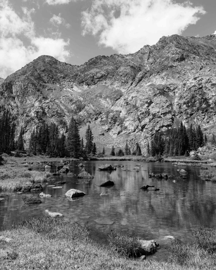 black and white image of mountains and trees with a lake reflection