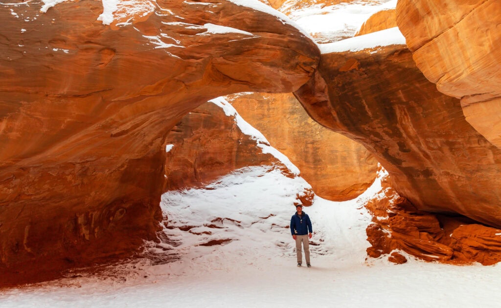 a hiker stands under sandstone arch with snow on the ground