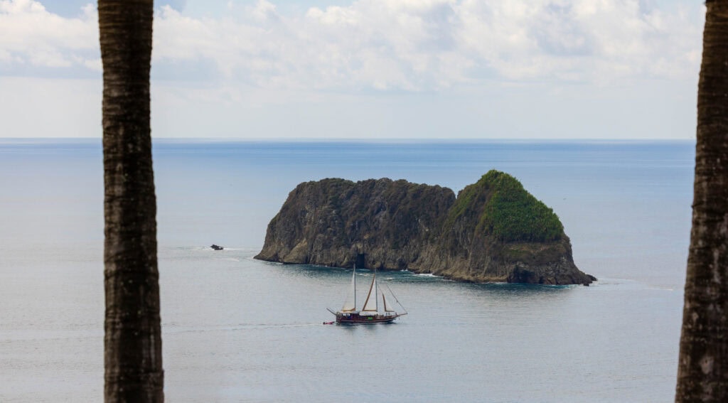 a wooden sail boat passes in front of a large rock island in costa rica