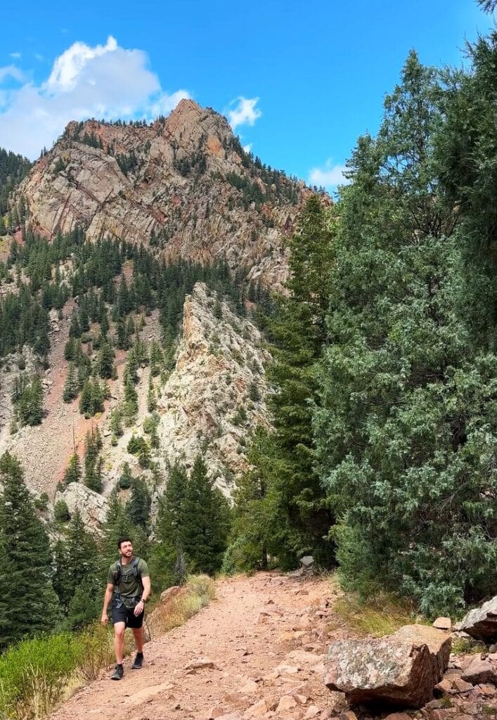 a male hiker in eldorado canyon state park with trees next to the trail and a big rock wall behind him
