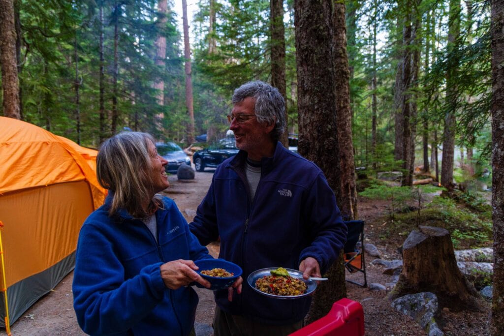 two people look at each other laughing while making food at a campground