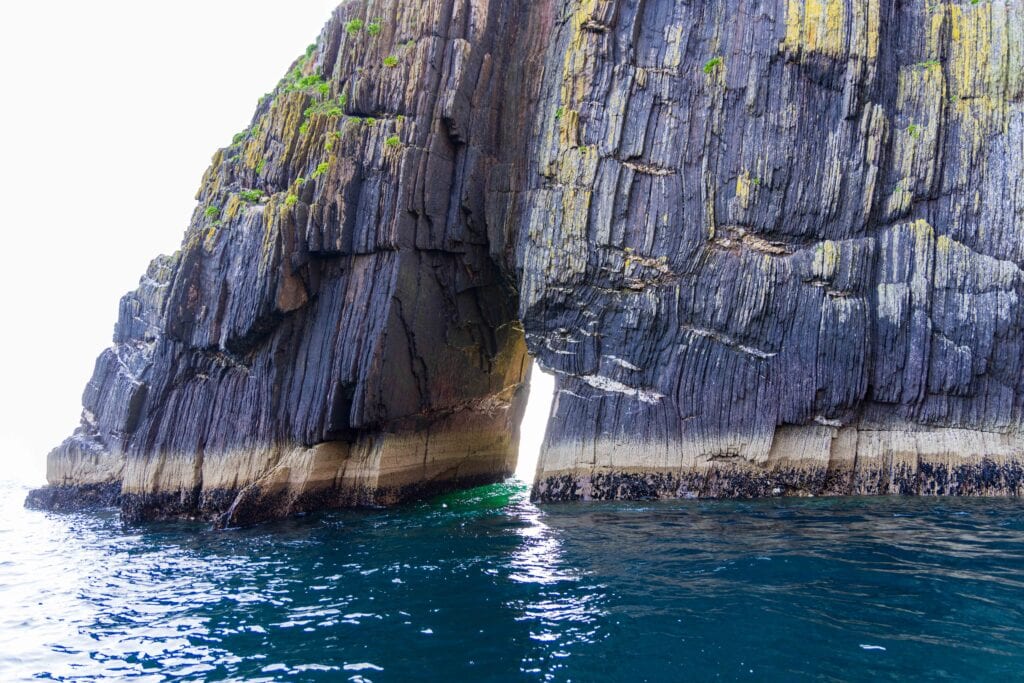 Rocky sea cliffs with a narrow natural arch opening along the Dingle coastline, viewed from a boat on a sea safari in Ireland.