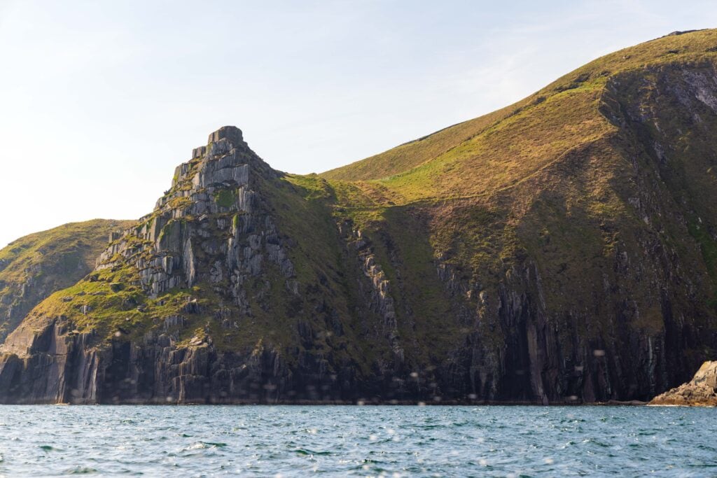 Wide view of rugged green-topped cliffs rising from the Atlantic Ocean along the Dingle Peninsula, seen on a boat tour in Ireland.