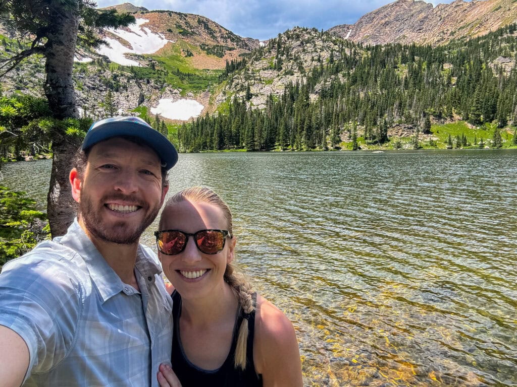 Couple taking a selfie at an alpine lake with forested mountains behind them