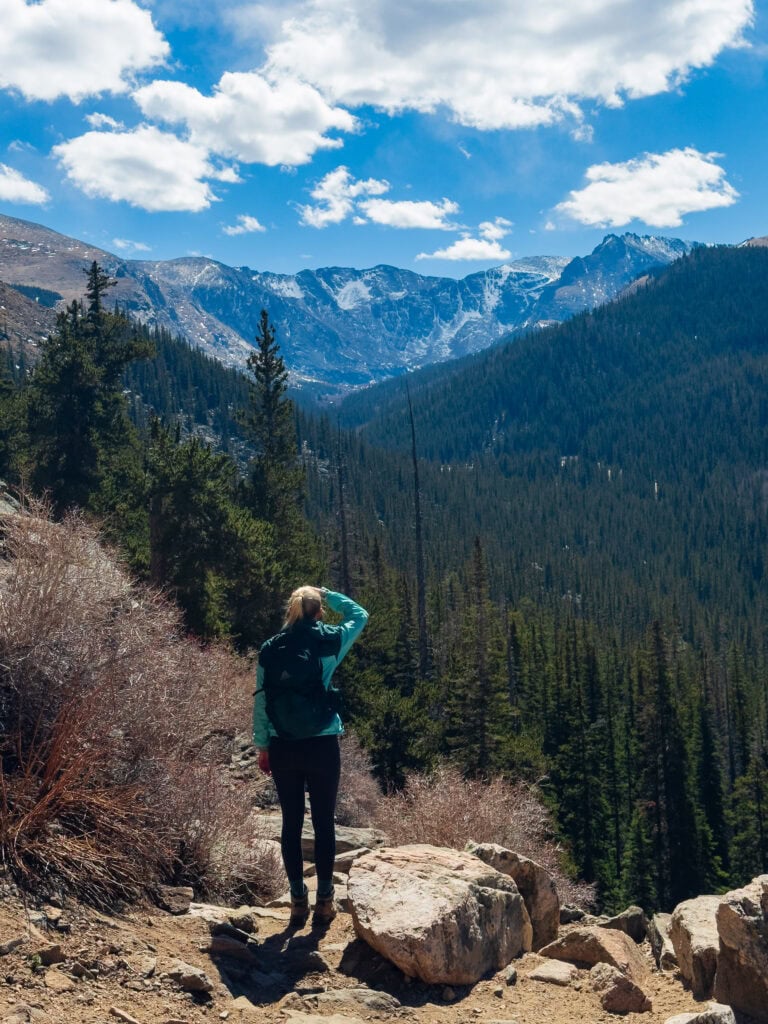 Hiker with backpack standing on a trail surrounded by pine trees with mountain peaks in the background
