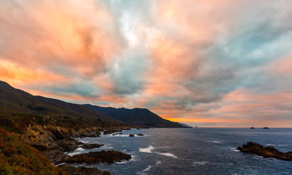 Warm pink and orange sunset clouds over a calm coastal bay with mountain silhouettes