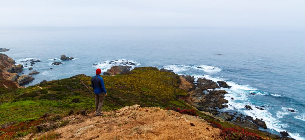 Hiker in a blue jacket standing on a clifftop overlooking the Pacific
