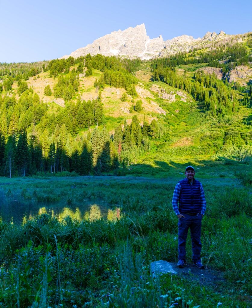 Hiker standing at the edge of a mountain lake with the Tetons in the background