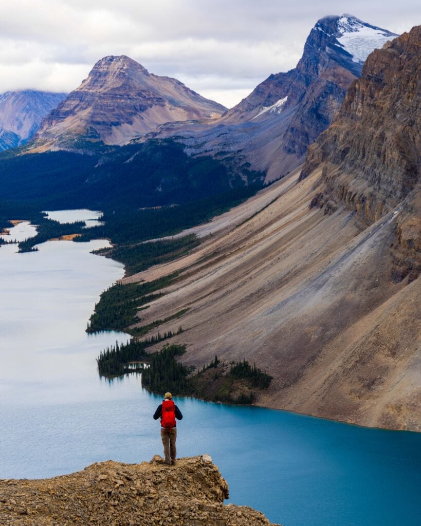 a single male hiker in a red backpack looks out over bow lake