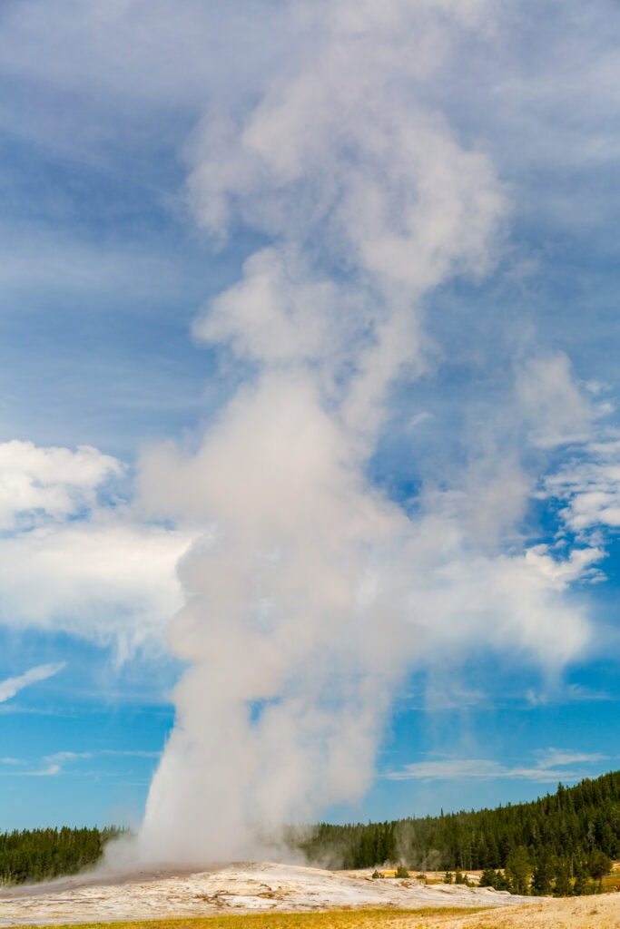 old faithful erupting with blue sky and clouds above