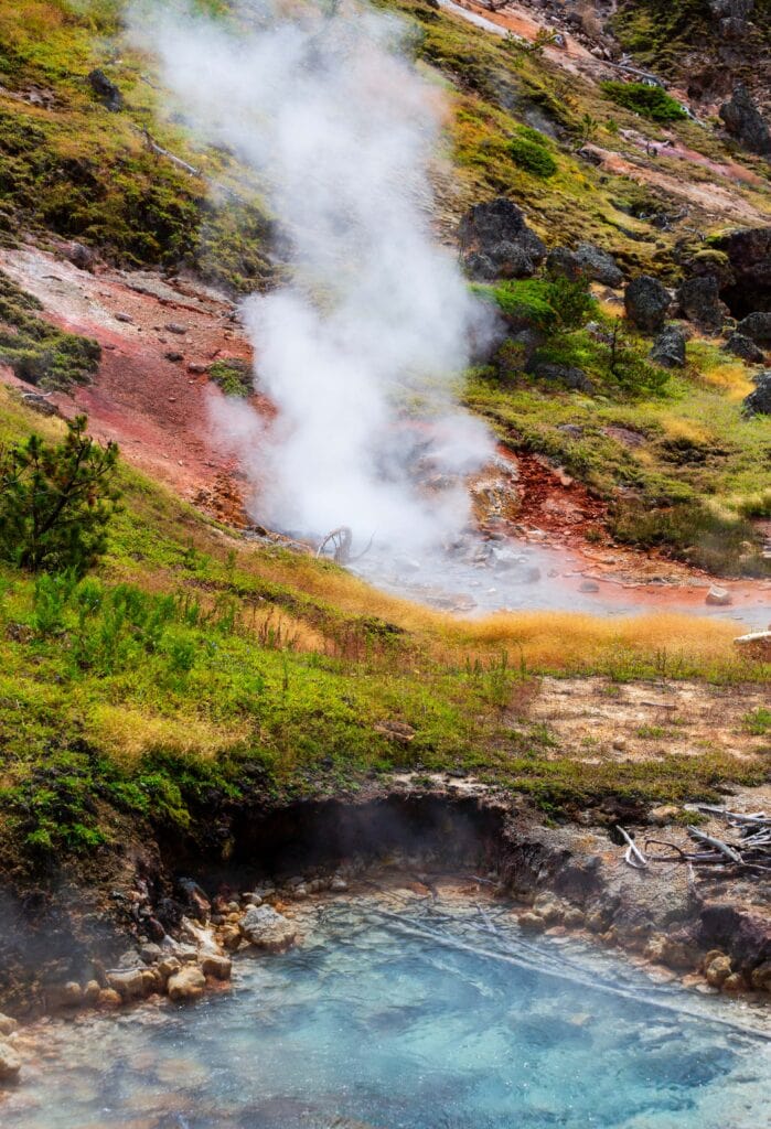 Geyser venting steam against a blue sky near a thermal hillside