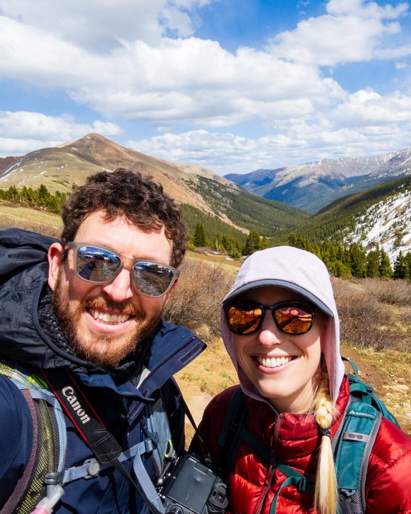 Smiling couple taking a selfie with mountain scenery behind them on Herman Gulch Trail in Colorado