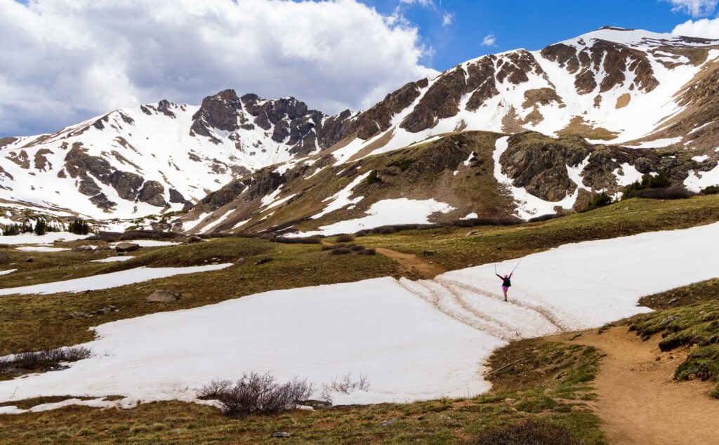 Hiker crossing a snowfield on Herman Gulch Trail with mountain peaks and scattered clouds in the distance