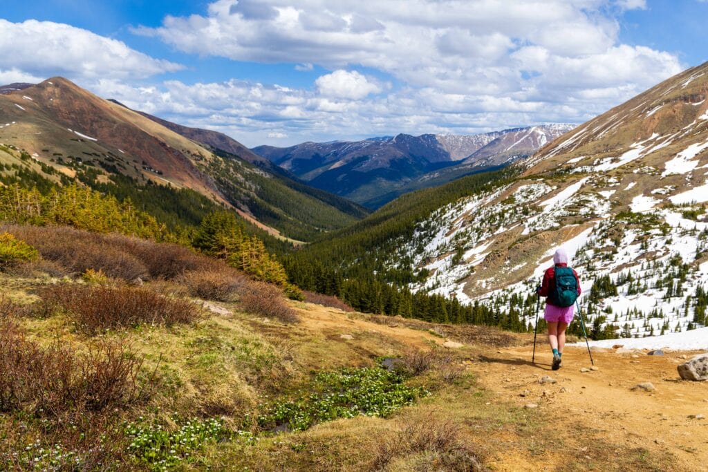 Hiker with a backpack descending a grassy trail toward a snow-streaked mountain ridge on Herman Gulch Trail in Colorado