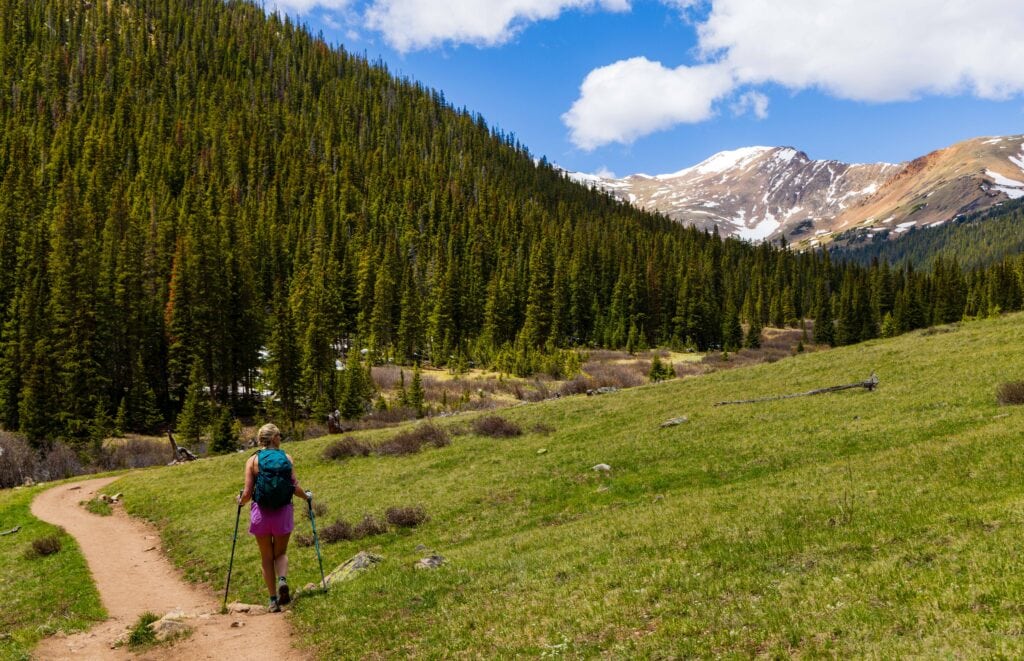 Hiker walking through a green alpine meadow on the Herman Gulch Trail with forested mountains in the background