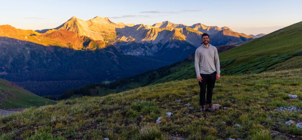 a hiker smiles with light hitting the mountain behind him