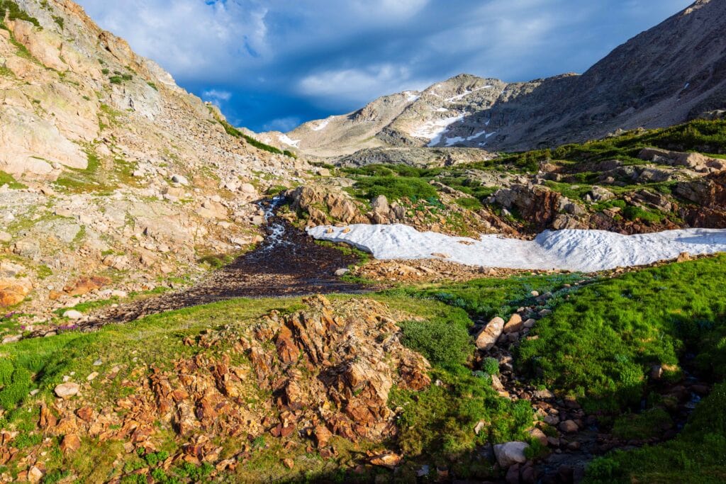 a lush basin with a creek flowing through it and some snow hanging on