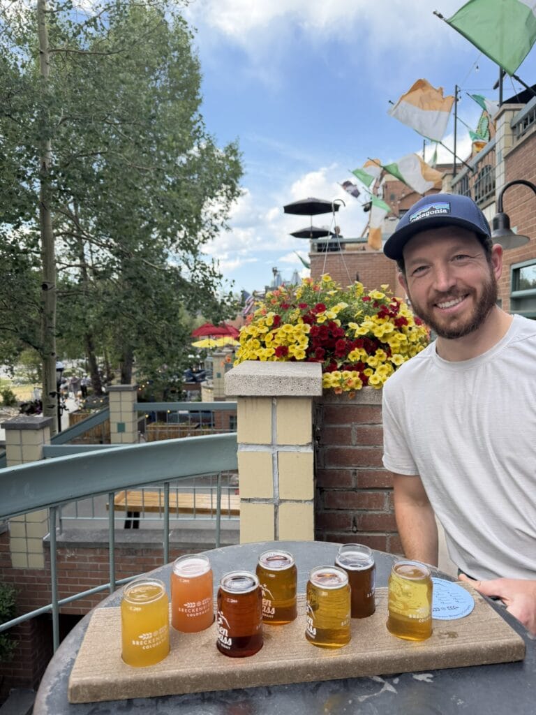a man smiles at the camera with a beer flight on the table