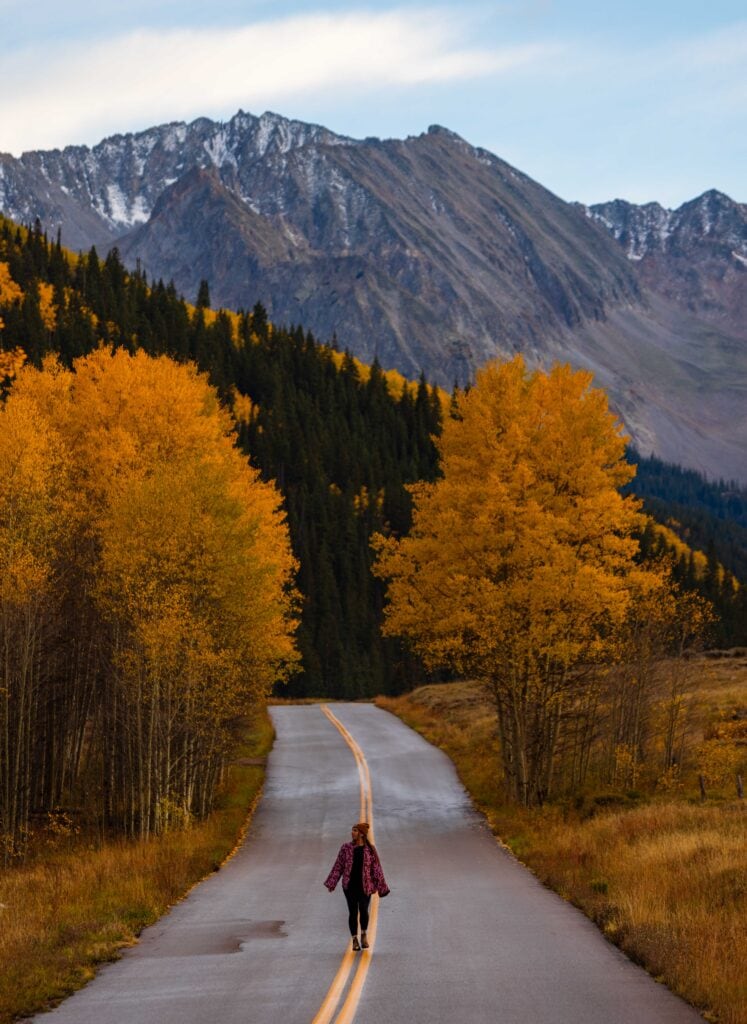 a woman walks down the middle of castle creek valley road with fall colors around her