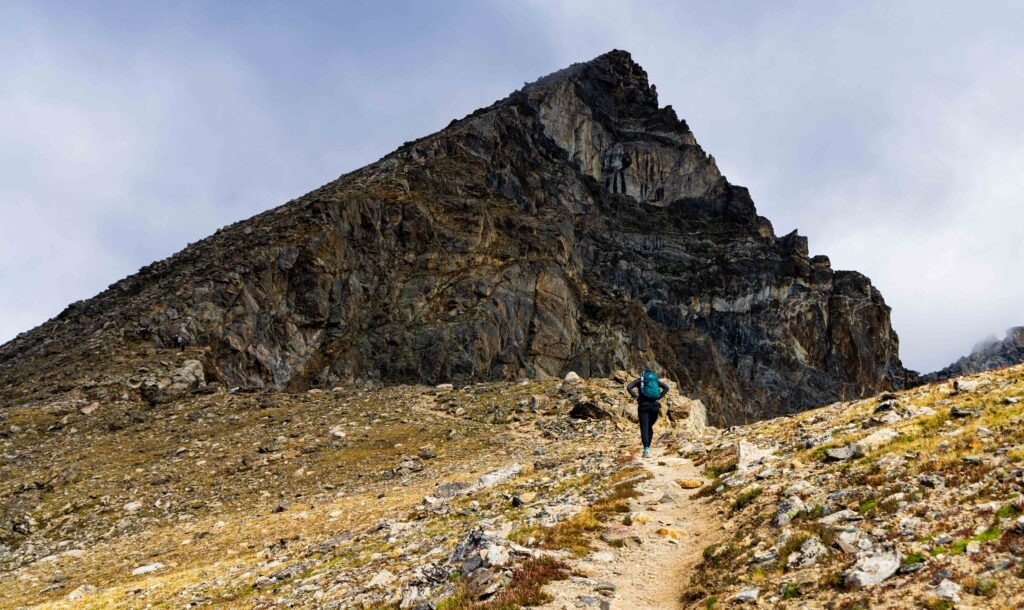 a hiker stands on the trail with a jagged peak above
