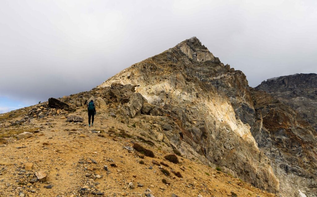 a hiker stands on the trail with a jagged peak above