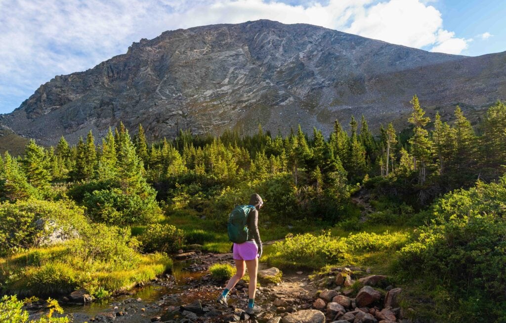 a female hiker crosses a small creek with south arapaho peak above her and trees next to her