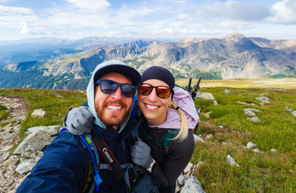 two hikers smile at the camera with lakes below