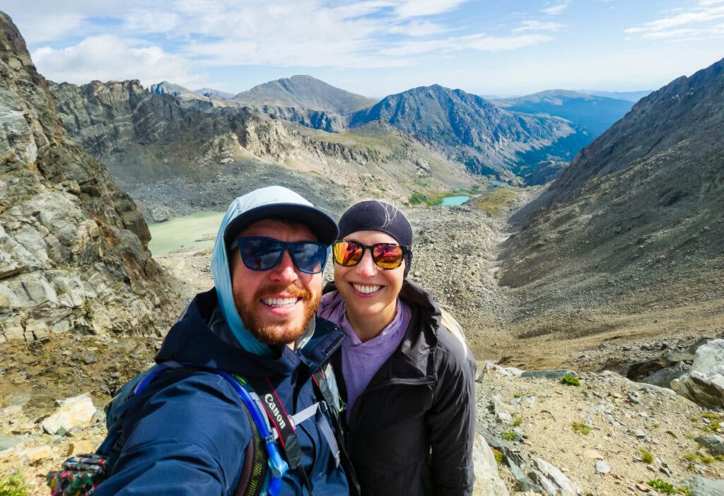 two hikers smile at the camera with lakes below