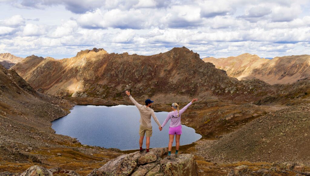 A couple standing on a rock with arms raised, overlooking an alpine lake